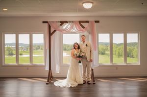 A couple stands in an elegant indoor venue with large windows overlooking a scenic landscape.