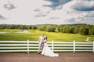 A couple poses along a white fence overlooking rolling green hills on their wedding day.