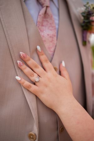 A close-up of a hand wearing an engagement ring against a tan suit jacket and pink tie.