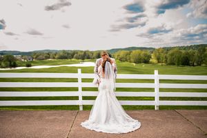 A bride in a lace gown stands against a white fence overlooking a scenic countryside landscape.