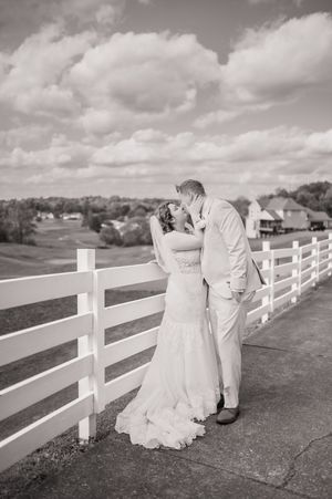 A black and white portrait of a couple embracing by a white fence with rolling hills in the background.
