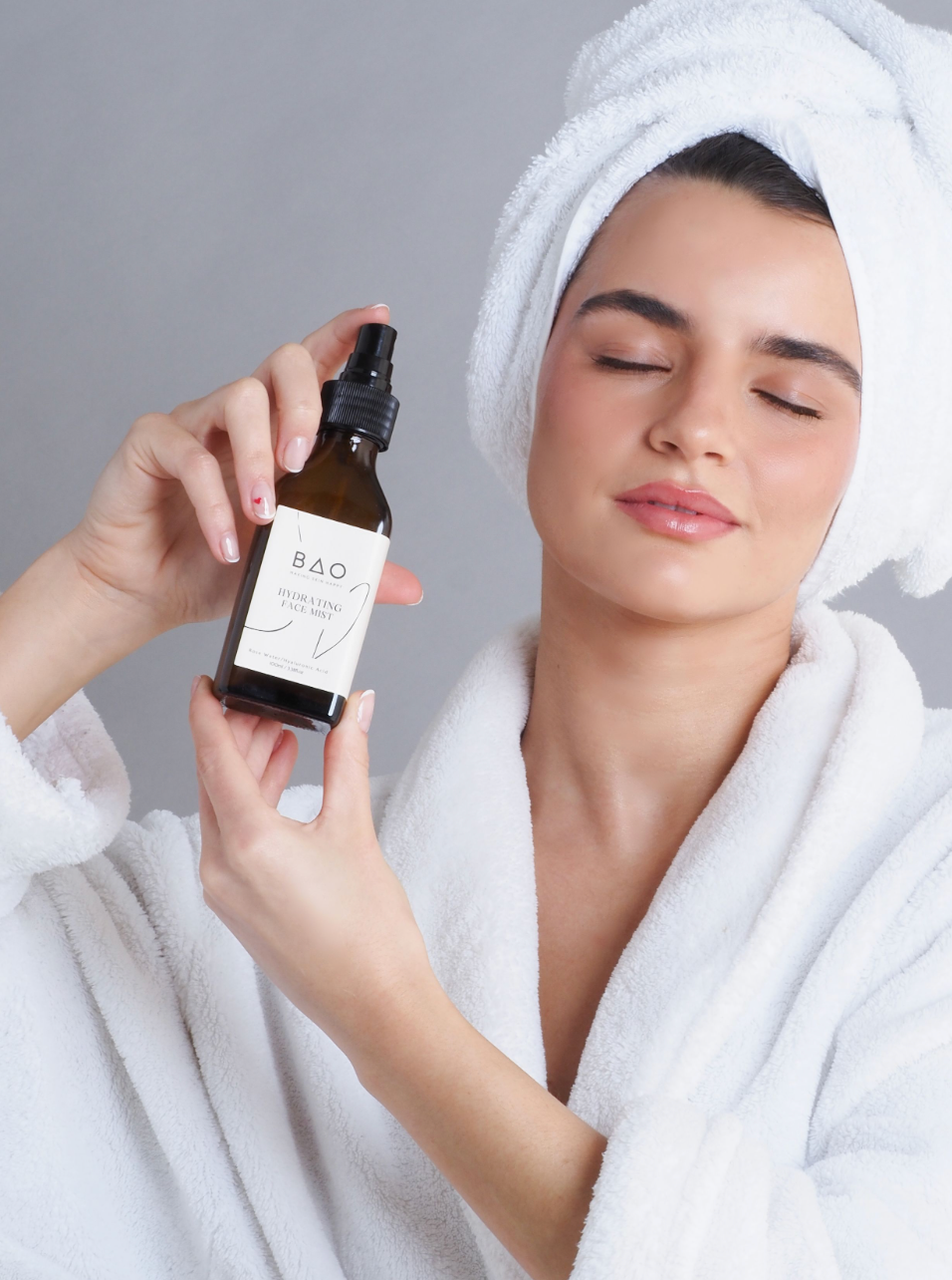 Someone wearing a white bathrobe and hair towel holds a RAO skincare product bottle while relaxing during a spa day.
