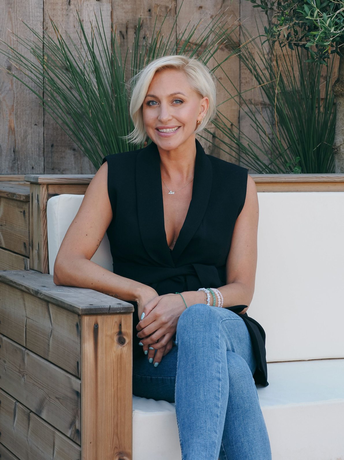 Casually dressed woman in black sleeveless top and blue jeans sitting on wooden bench with ornamental grass background.