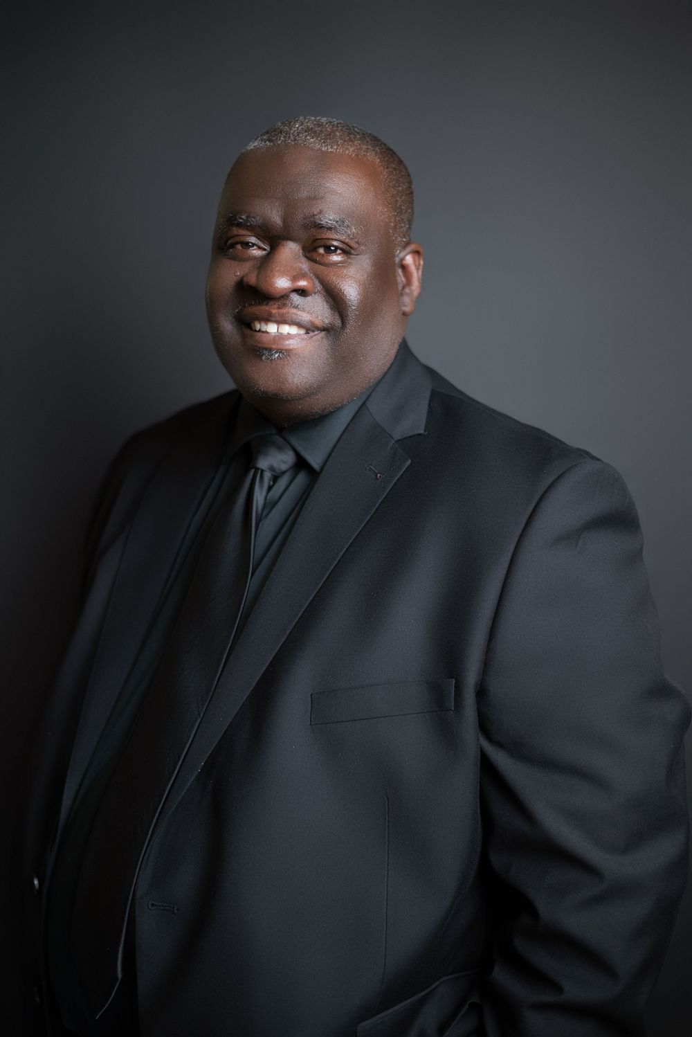 Professional headshots of person in black suit against dark studio background.