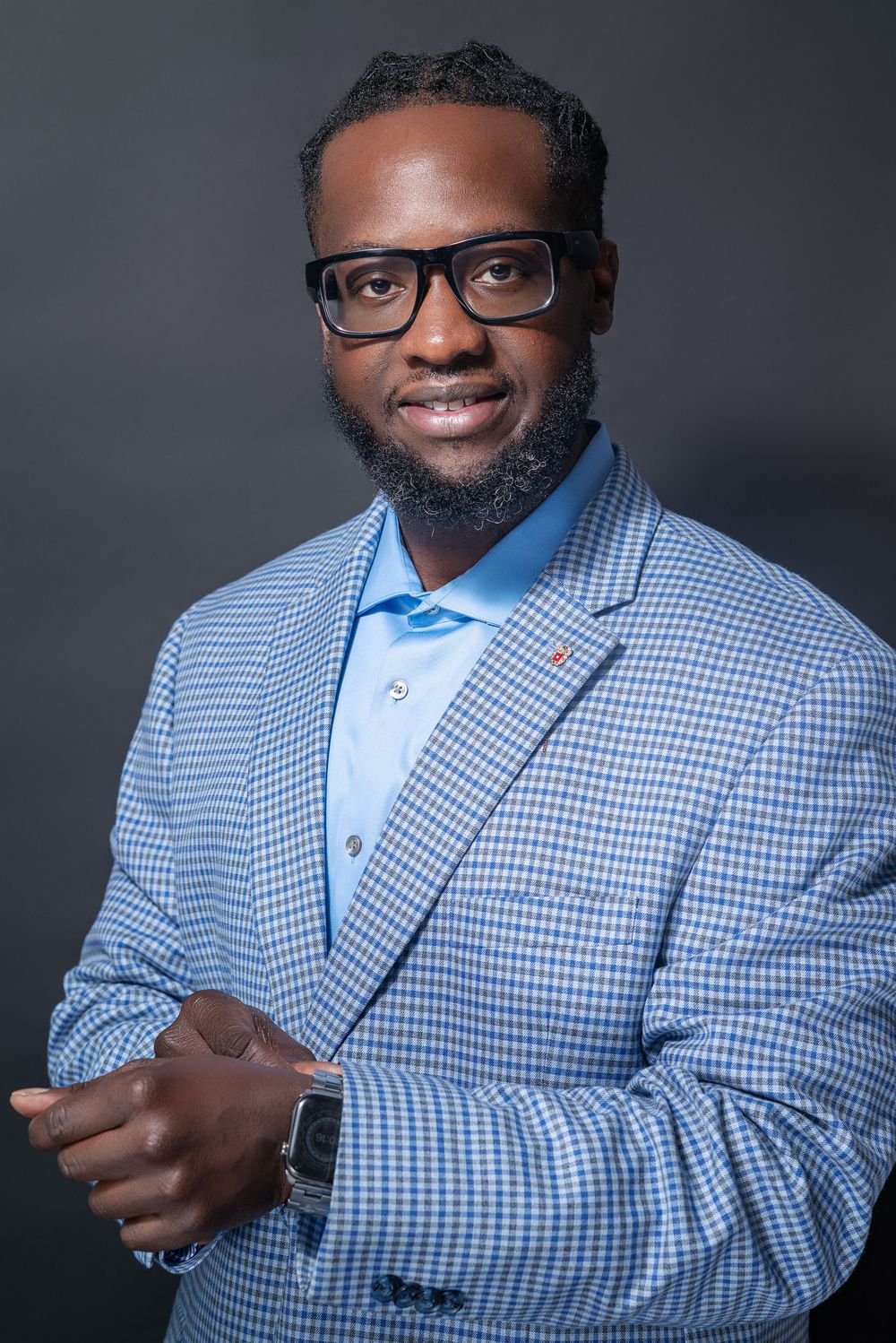Professional headshot of a person wearing glasses, a light blue dress shirt and checkered blazer against a dark background.