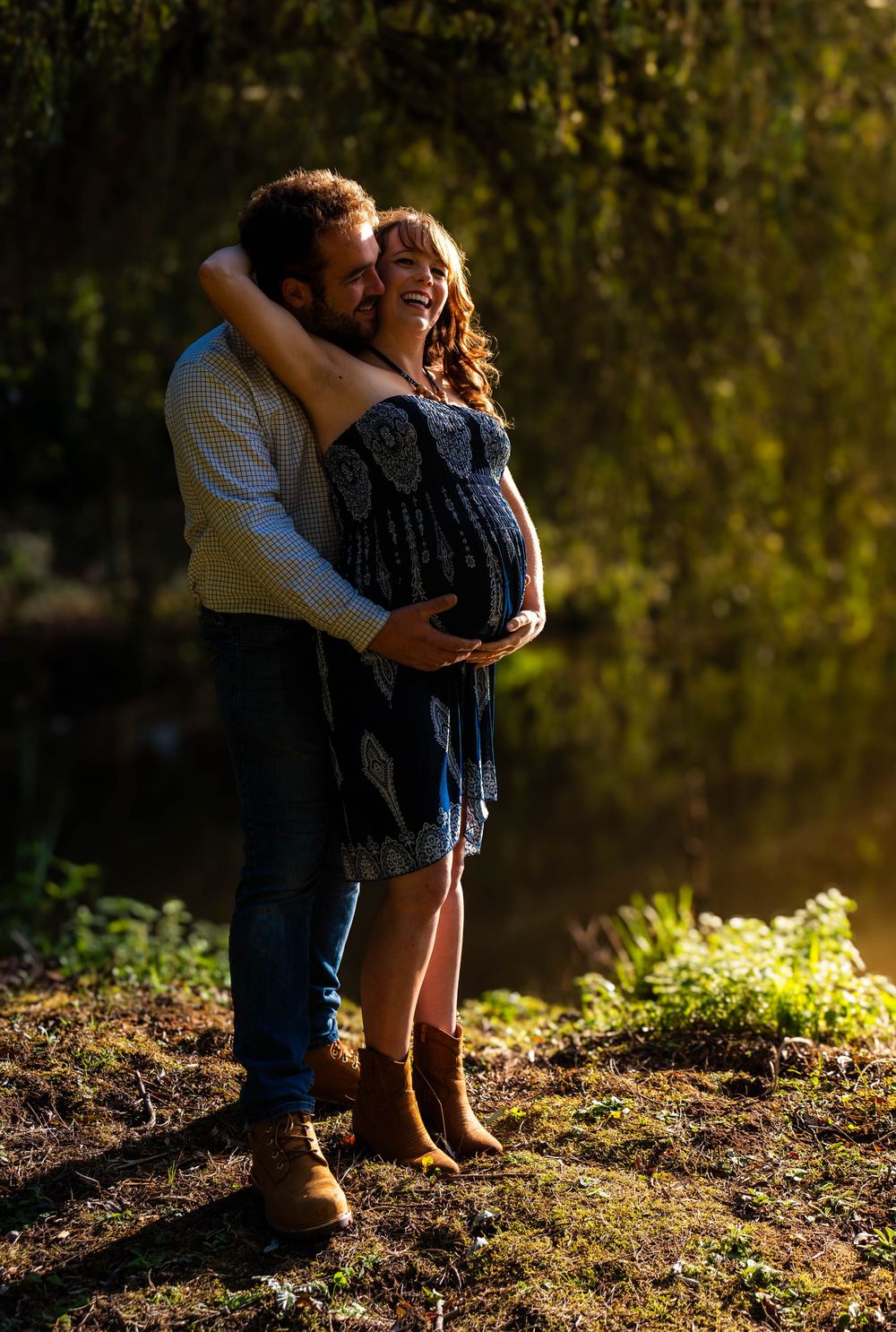 A tender maternity moment captured in golden sunlight near a tree-lined shore.