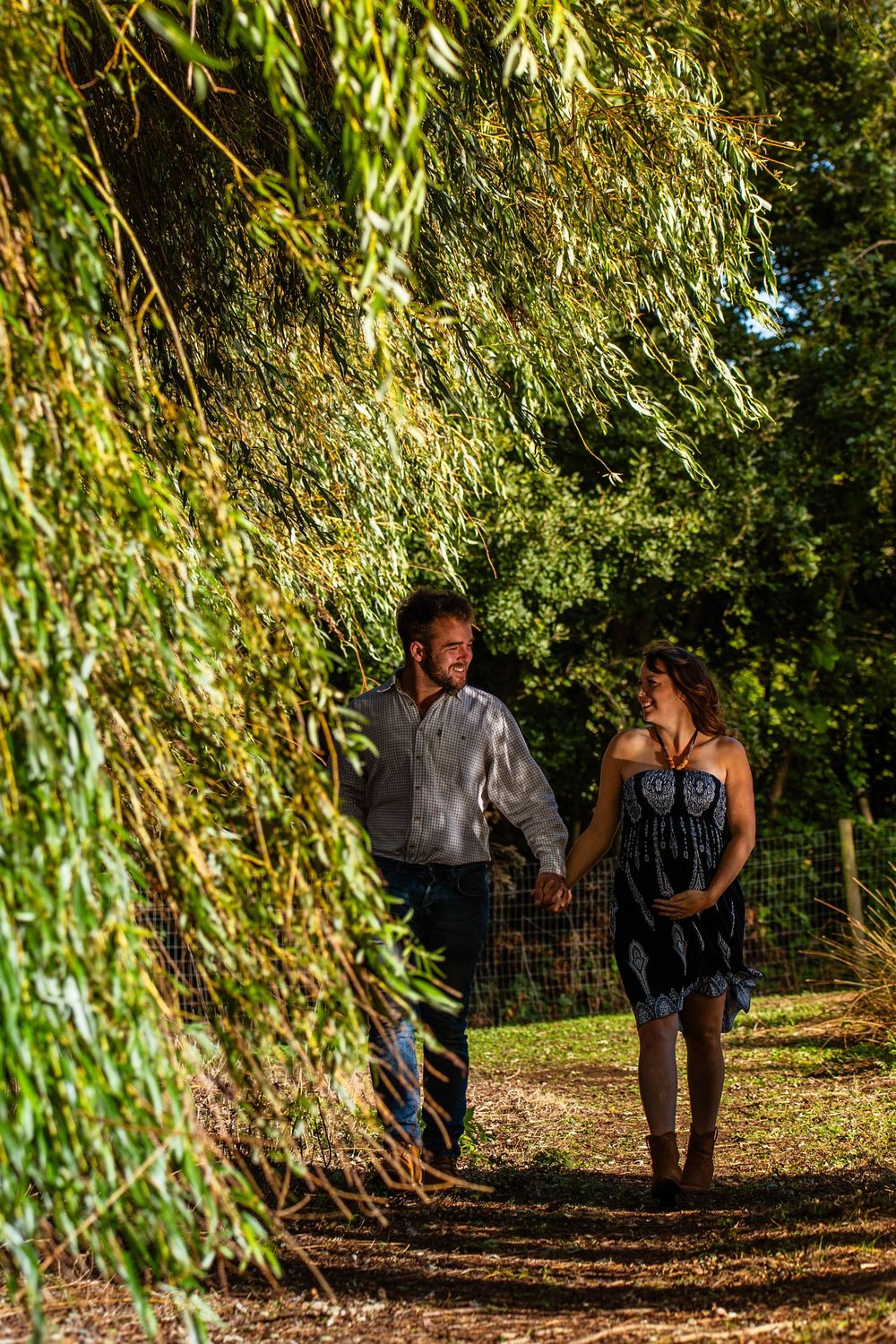 A couple walks hand in hand under draping willow trees on a garden path.