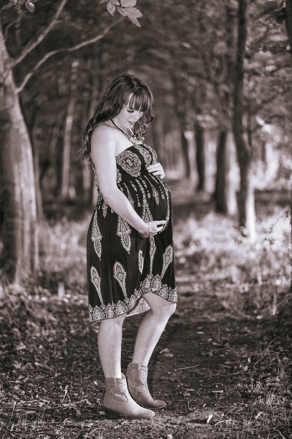 A serene maternity portrait in black and white showing a person in a patterned dress among trees.