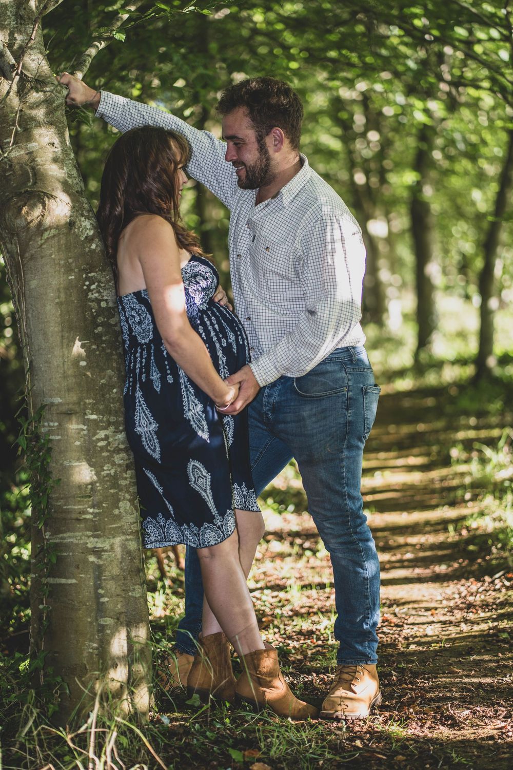 A romantic maternity photo of a couple embracing against a tree in dappled sunlight.