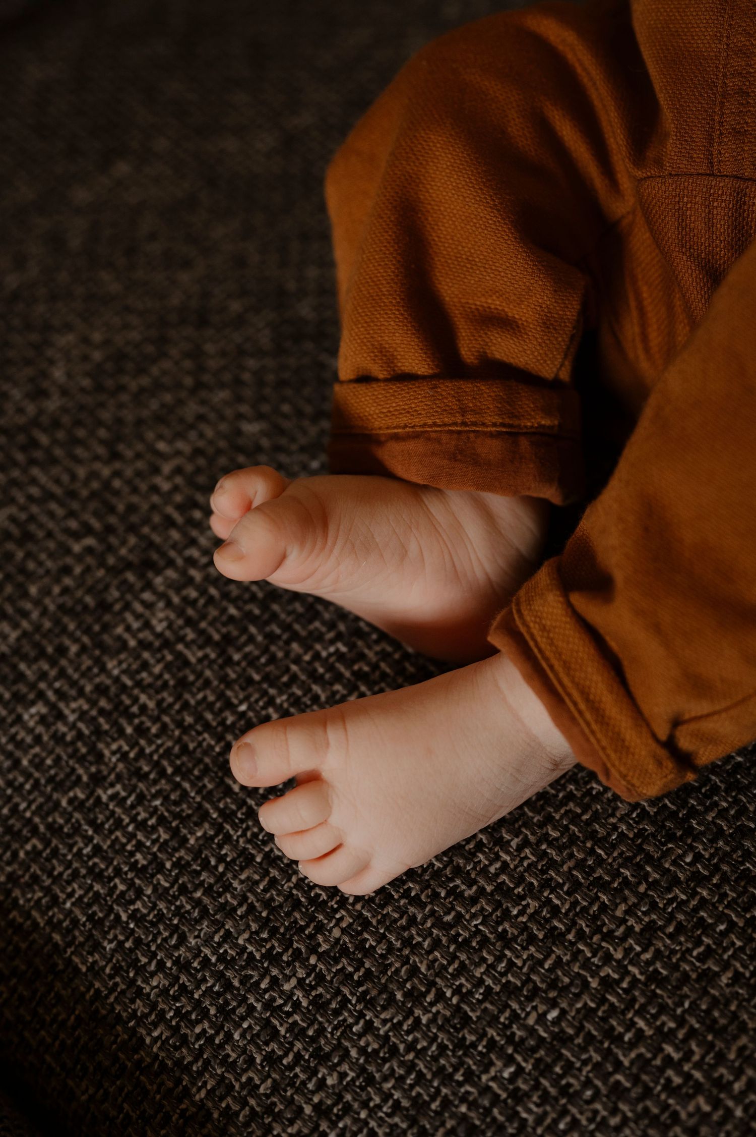 Close-up view of tiny barefoot baby feet against dark textured fabric with brown clothing visible.