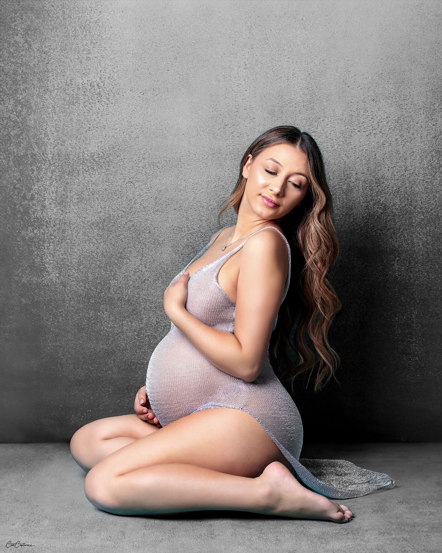 pregnant woman sitting on floor of gray canvas looking stunning in silver net dress