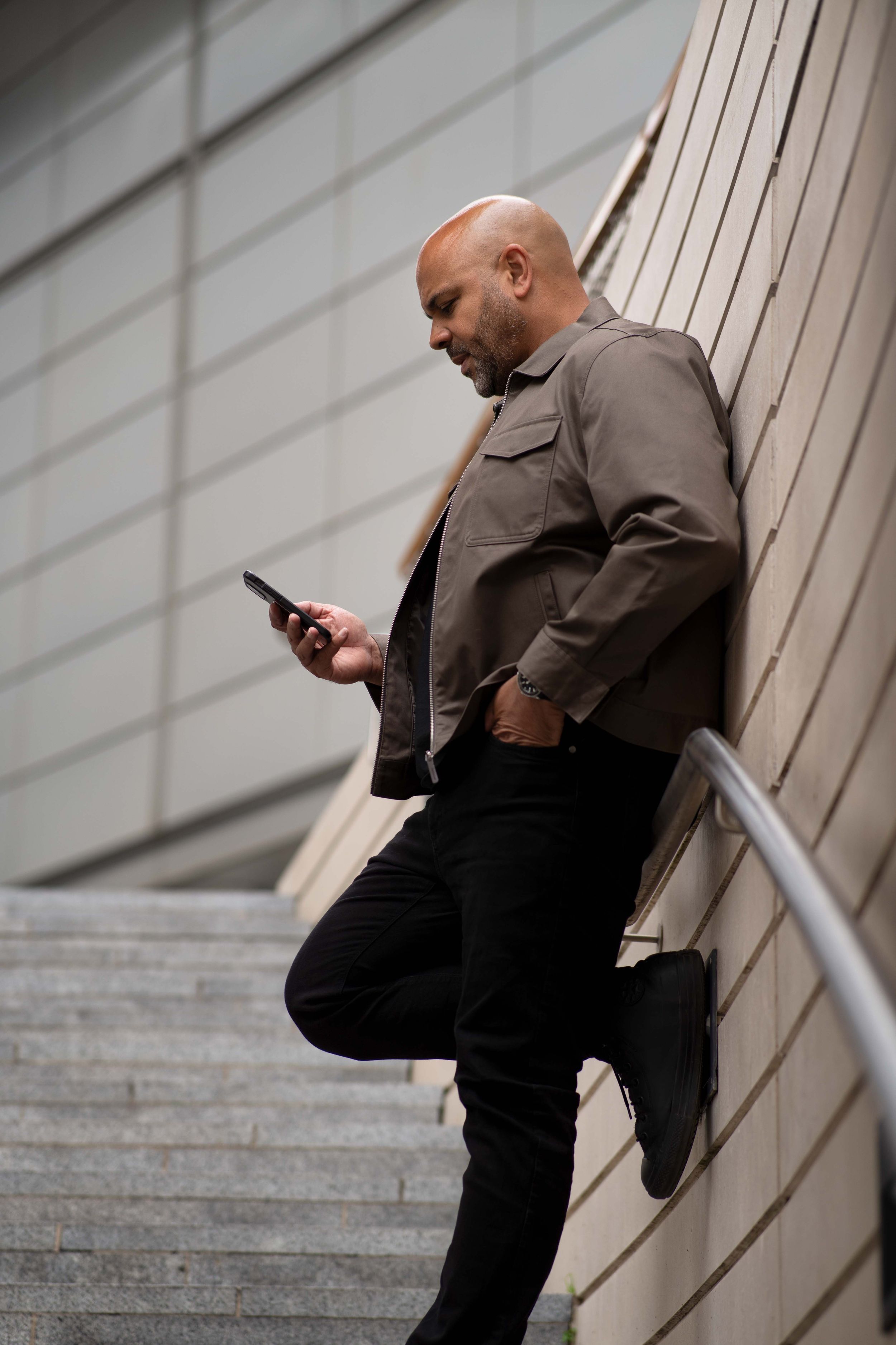 Person in brown jacket and black pants casually checking phone while leaning against stairway railing.