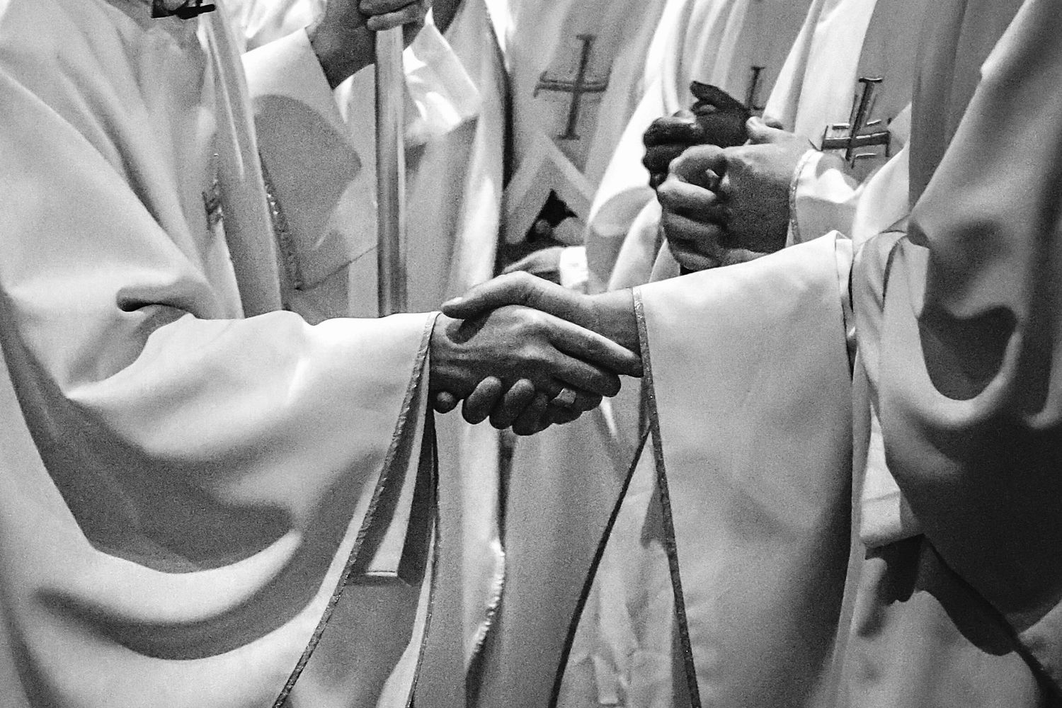 A Catholic Bishop and a priest wearing white ceremonial robes grasp hands in formal greeting.