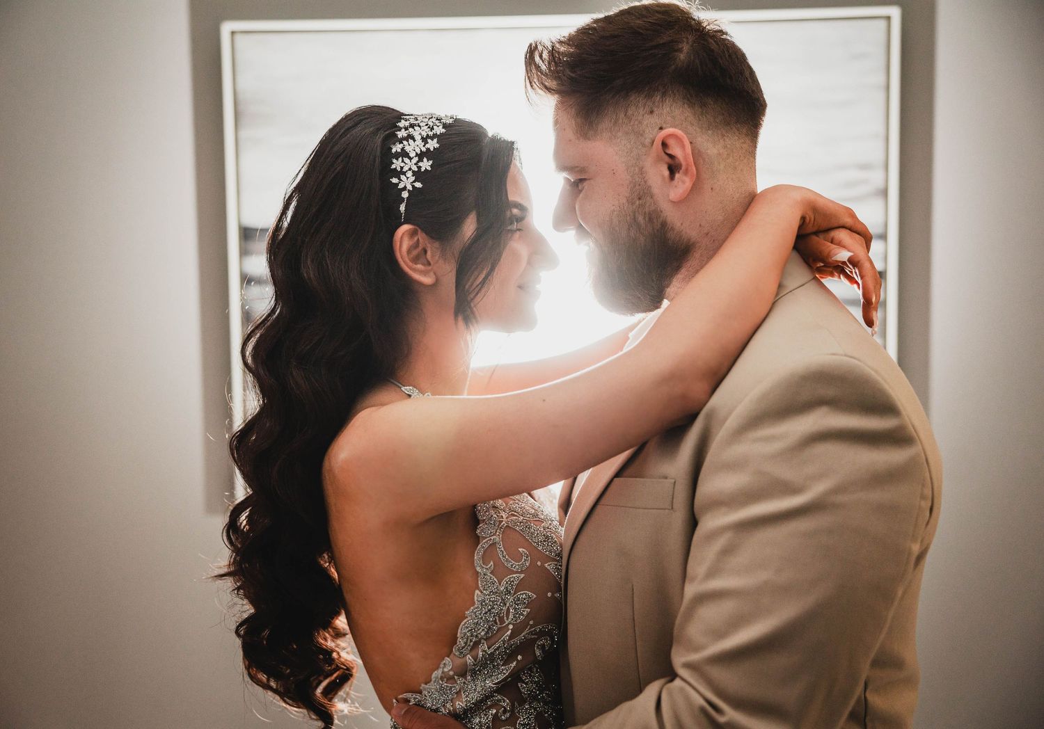 A romantic wedding couple embraces in soft window light with the bride wearing a crystal hairpiece and flowing dark hair.
