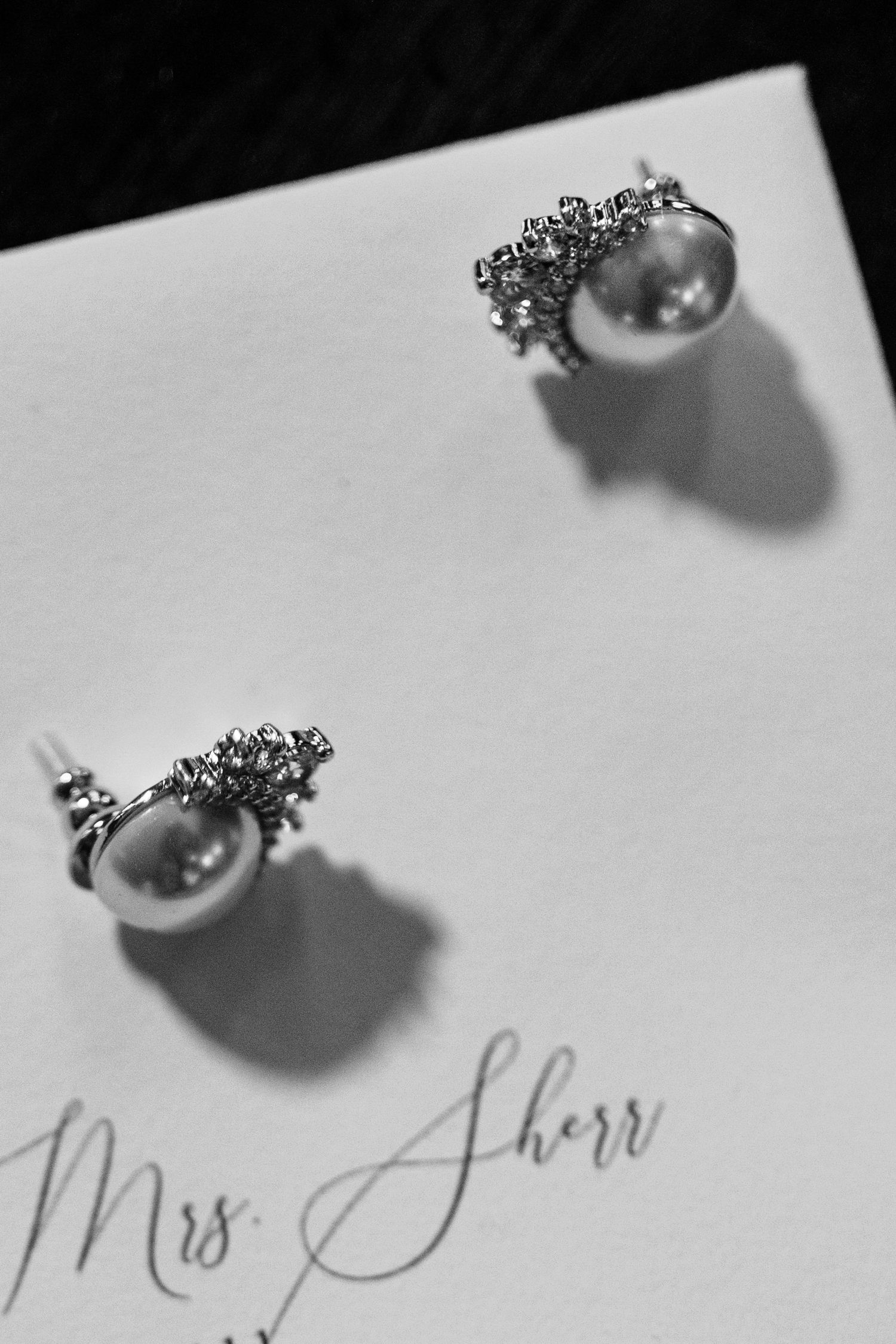 Elegant pearl and diamond stud earrings cast dramatic shadows on white paper with cursive text reading 'Miss Sherr'.