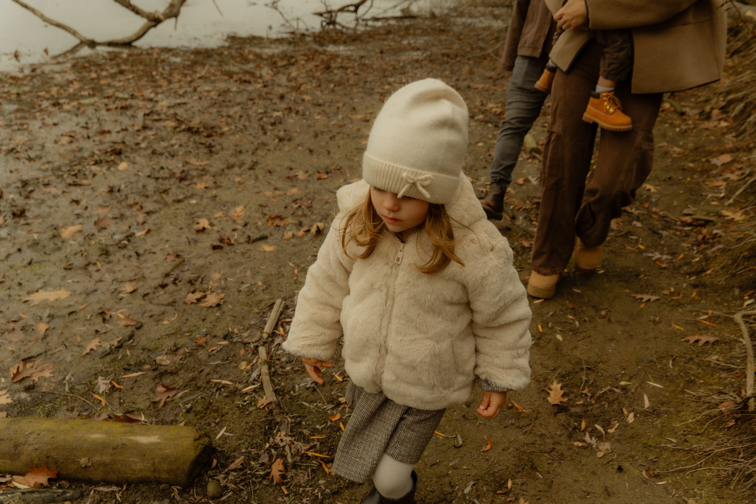 A small figure in a cream-colored winter coat and beanie walks along a muddy path on an autumn day.