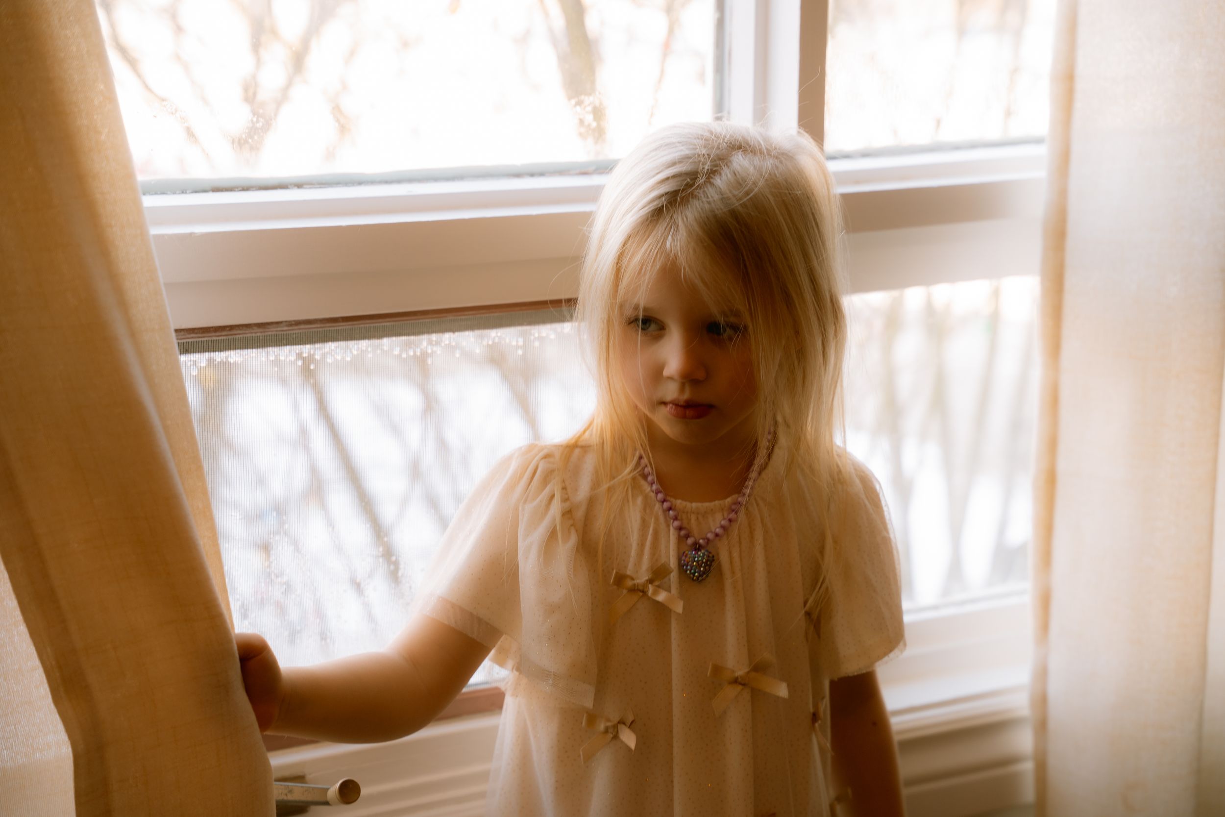 Soft dreamy vintage photo of someone in a white dress standing by a sunlit window with sheer curtains.