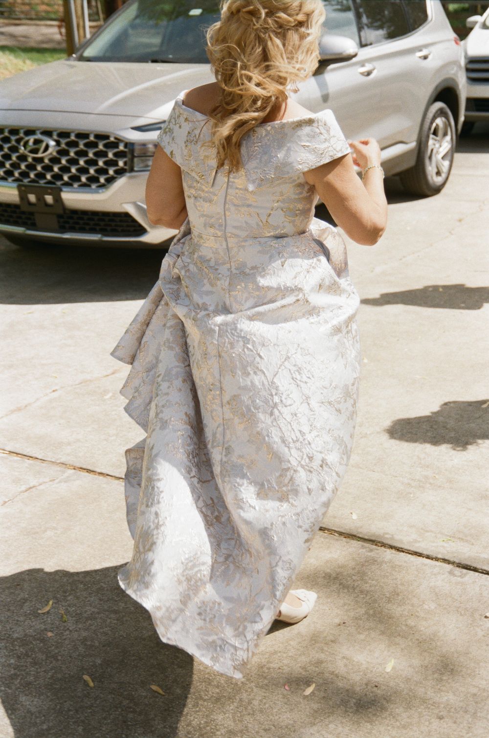 Back view of a bride in an elegant white floral brocade wedding dress with cap sleeves walking on a sidewalk.