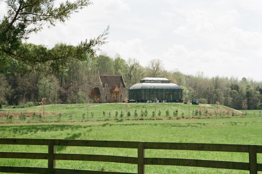 Rural farmland with wooden fence, rolling green pastures and rustic barn buildings in the background.
