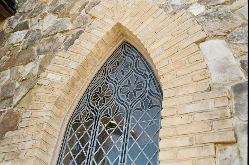 Detail of an ornate Gothic church window with diamond-patterned iron grating set within a tan stone archway.