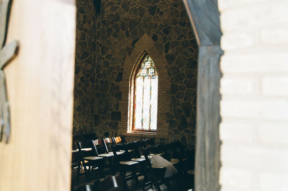 View through a gothic stone archway showing stained glass window and wooden pews in a dimly lit church interior.