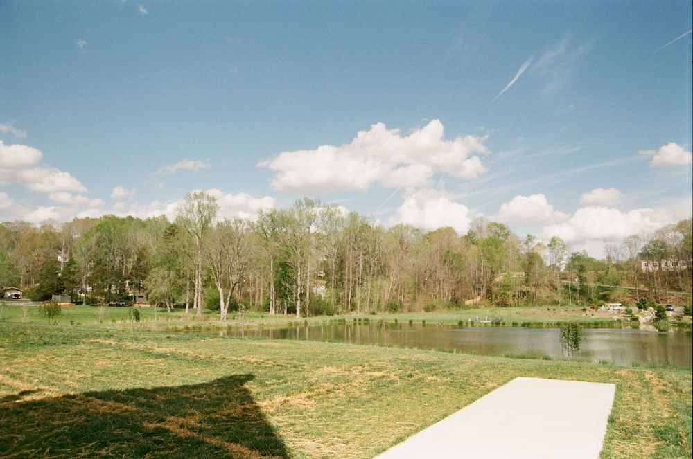 A scenic landscape view of a pond surrounded by trees and grassy fields with a white path leading to the water's edge.