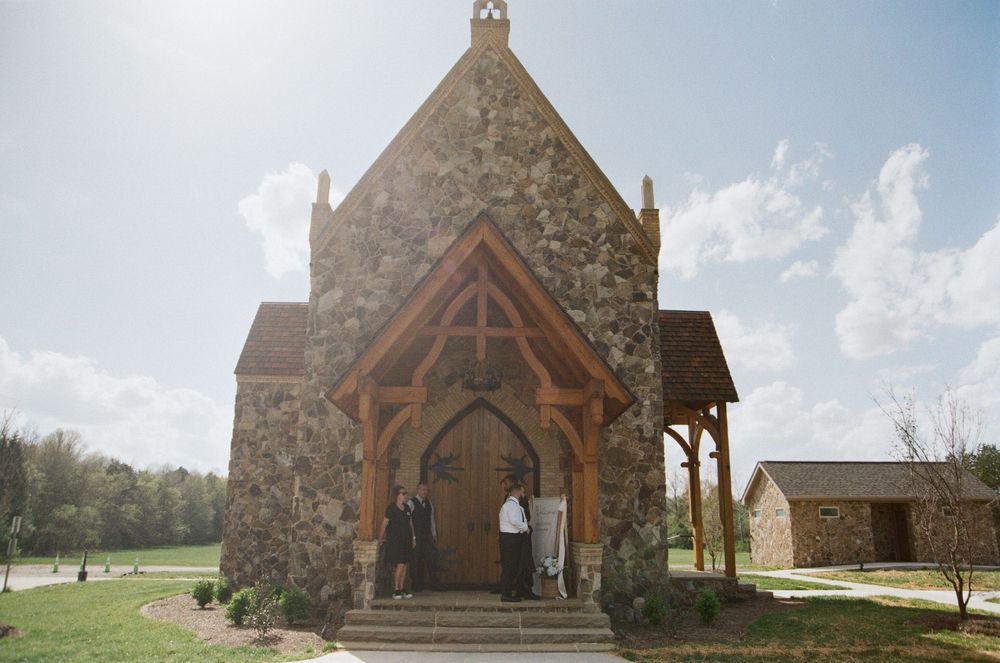 Rustic stone chapel with wooden archway entrance and sunlight shining behind its steeple on a bright day.
