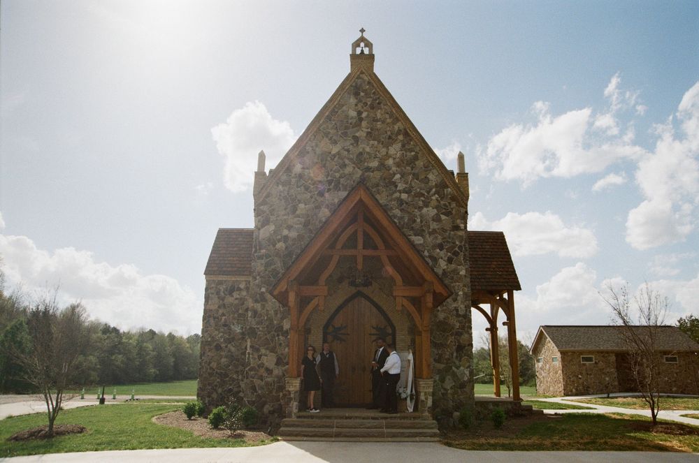 Small stone church with wooden portico entrance and pointed arch doorway stands against blue sky on sunny day.