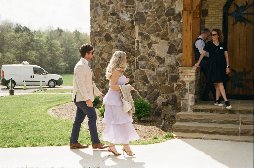 A couple walks toward a stone building entrance where two people are standing at the doorway on a sunny day.