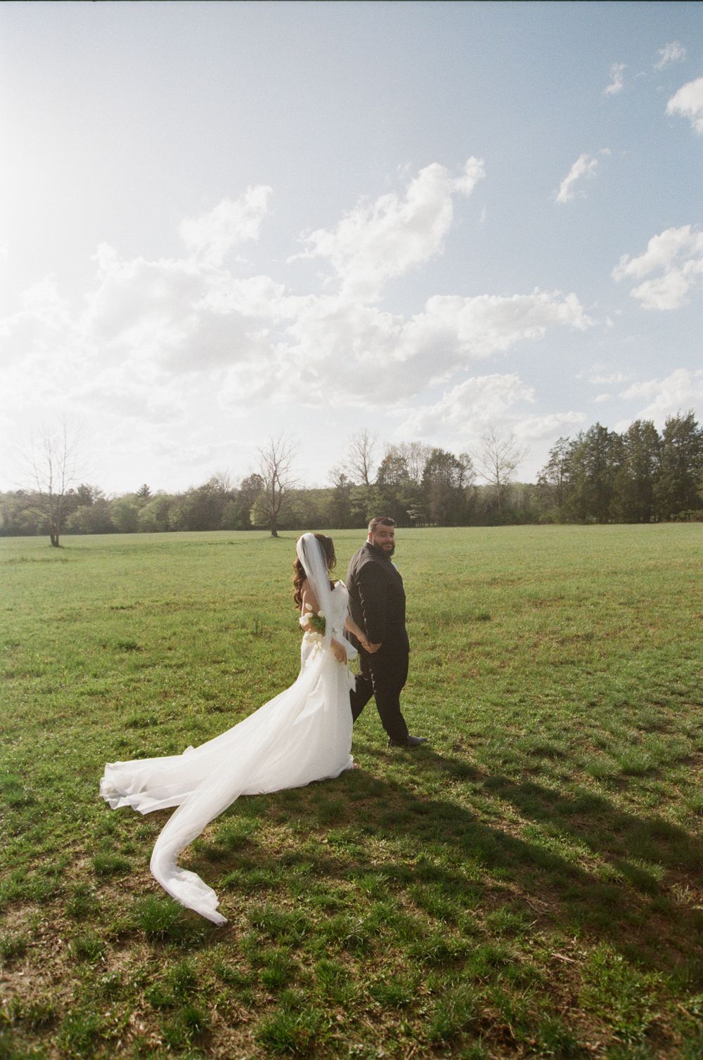 A couple in wedding attire walks through a sunlit green field on their special day as clouds drift overhead.