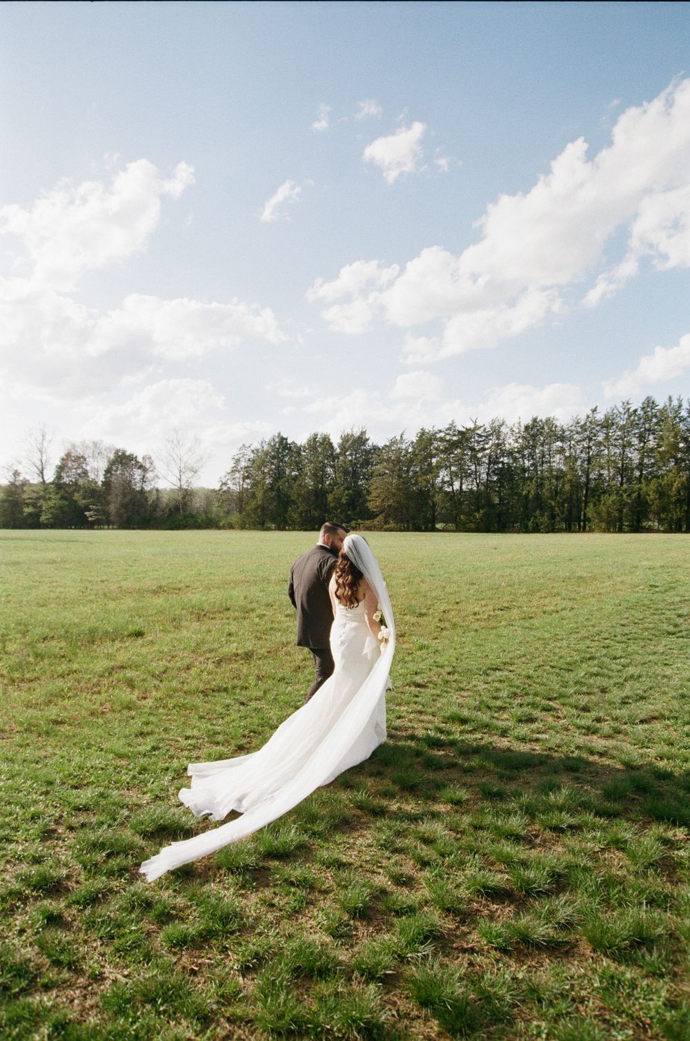A couple shares an intimate moment in a green field with flowing white wedding dress and treeline in the background.