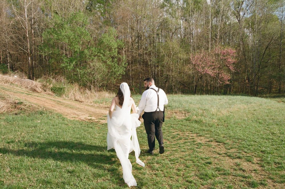A couple in wedding attire walks up a grassy hill path surrounded by trees on a sunny day.
