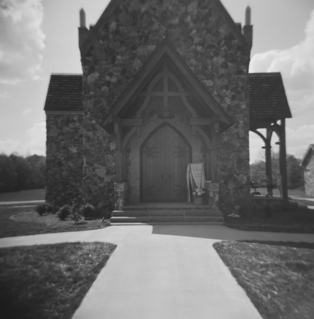 Black and white photograph of a stone church entrance with arched doorway and concrete path leading to wooden doors.