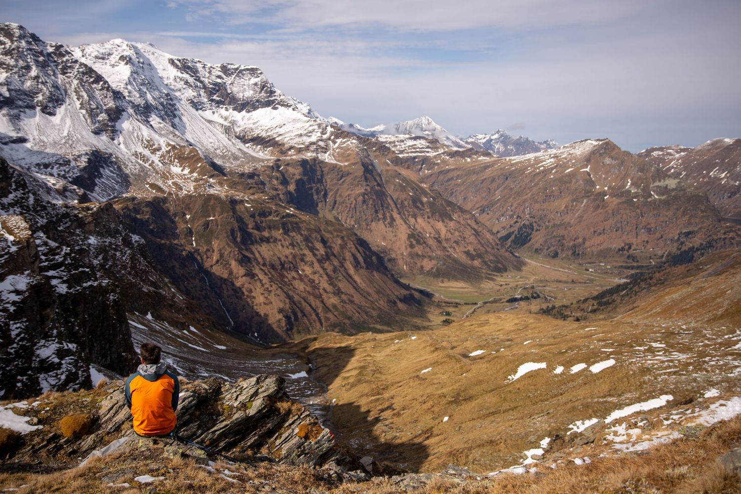 Hiker in orange jacket sits on mountain ledge overlooking snow-capped peaks and dramatic valley below.