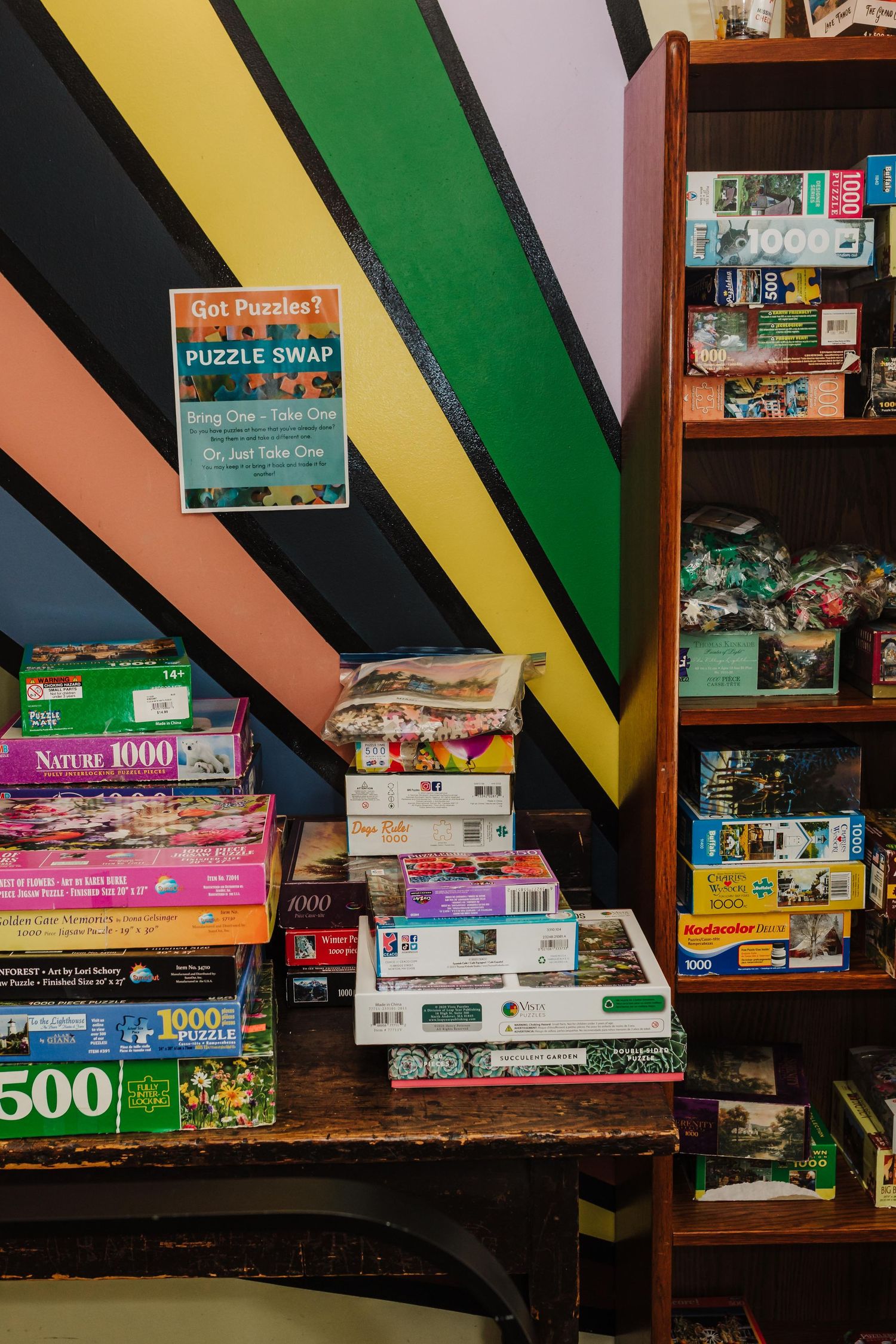 Stacks of puzzle boxes arranged against a colorful striped wall with a wooden bookshelf with items for Myerstown Library's Puzzle Swap.