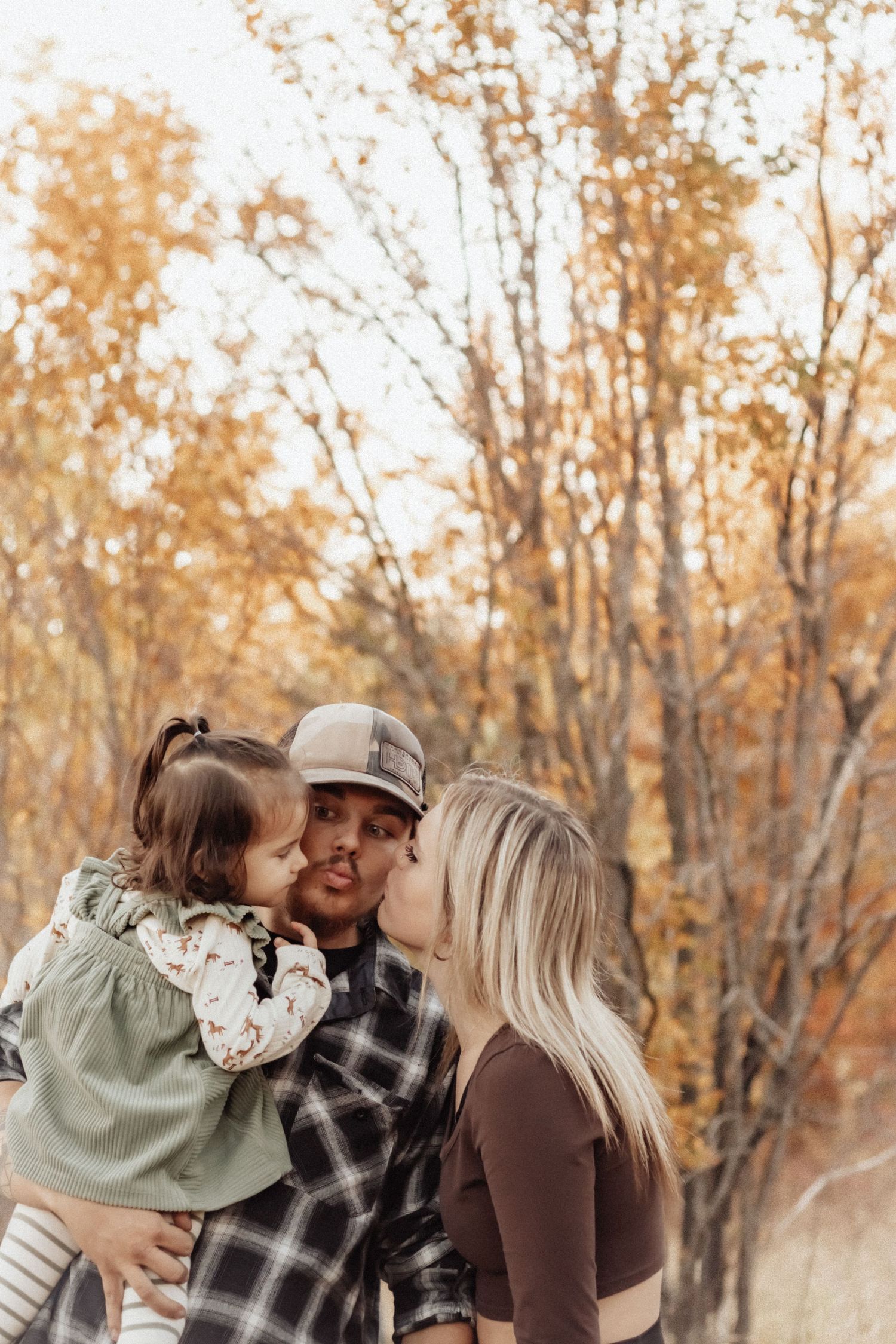 Family sharing a kiss together against autumn trees in warm lighting.