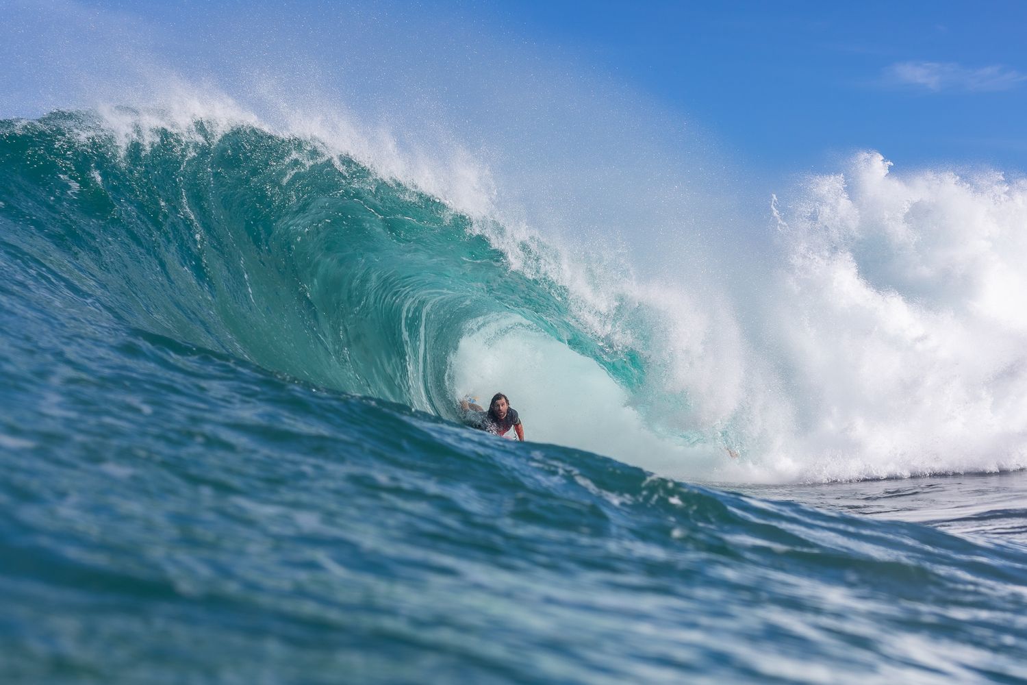 A surfer rides inside a massive turquoise wave barrel at a tropical surf break on a sunny day.