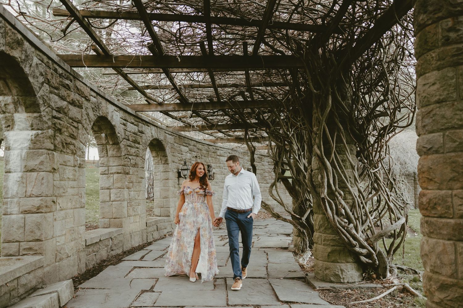 A romantic couple walks hand in hand under a stone archway covered in twisted vines during an outdoor photoshoot.