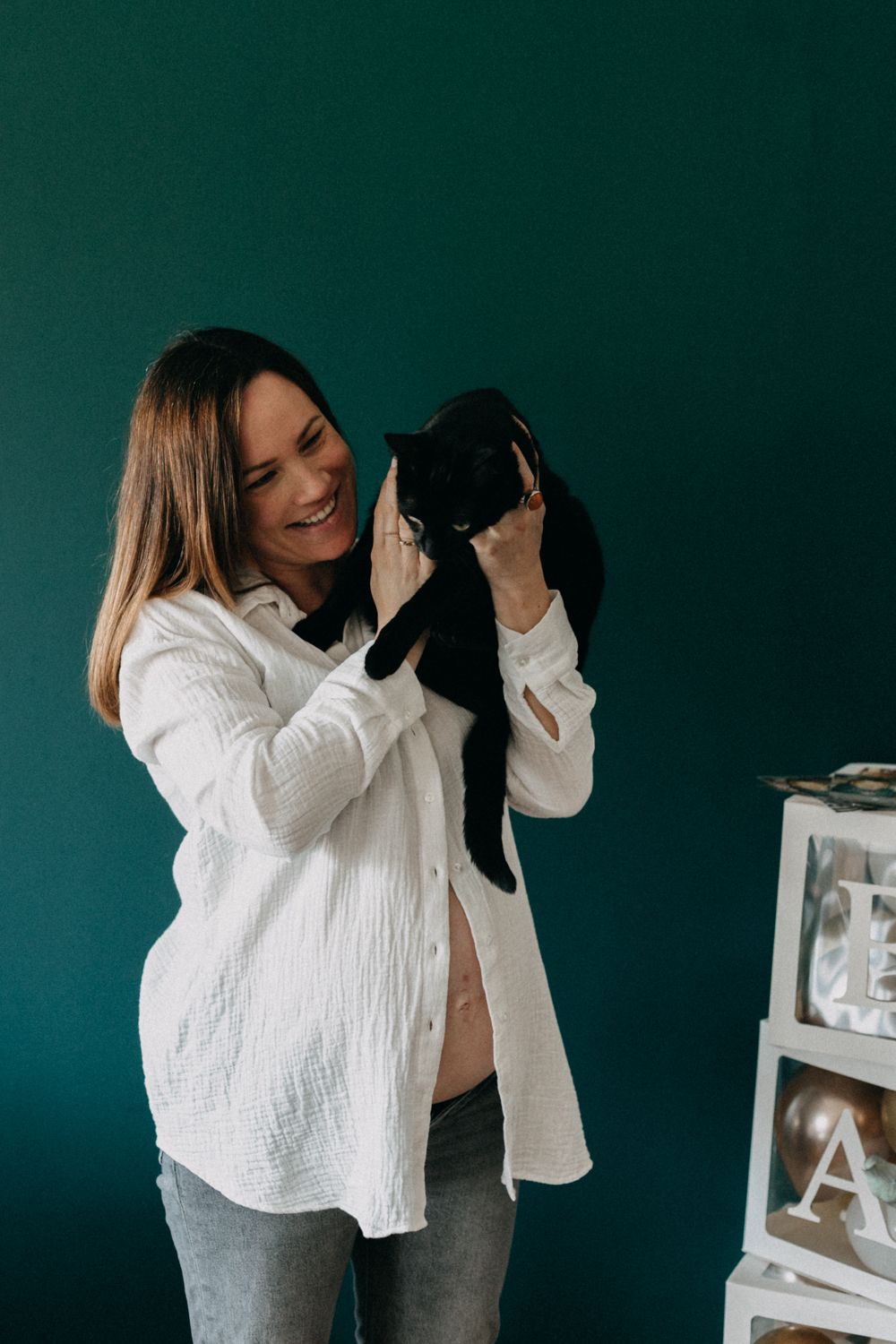 A smiling person in a white shirt holds and plays with a black cat against a dark teal wall.