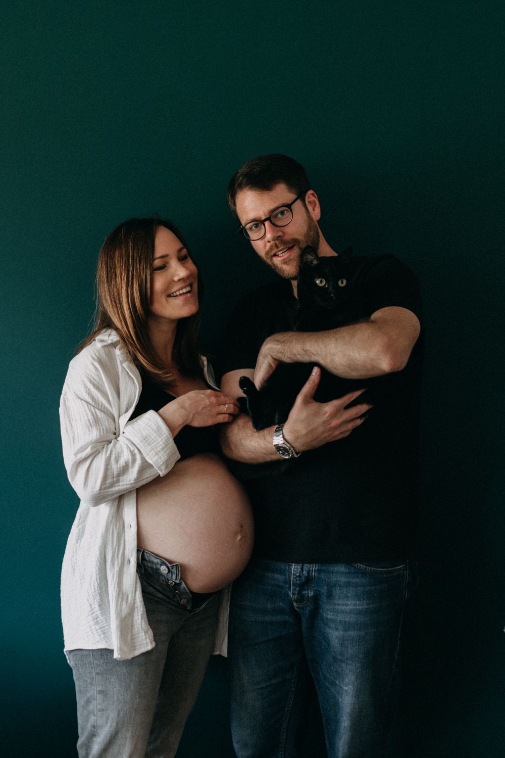 A maternity photo featuring a couple and their black cat against a dark green backdrop.