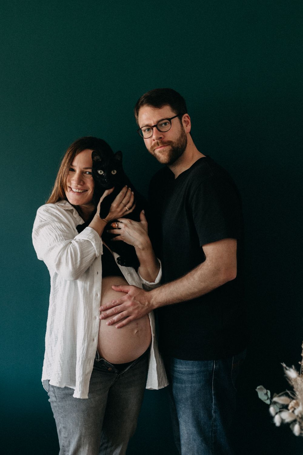 An expecting couple poses with their black and white cat against an emerald green background.