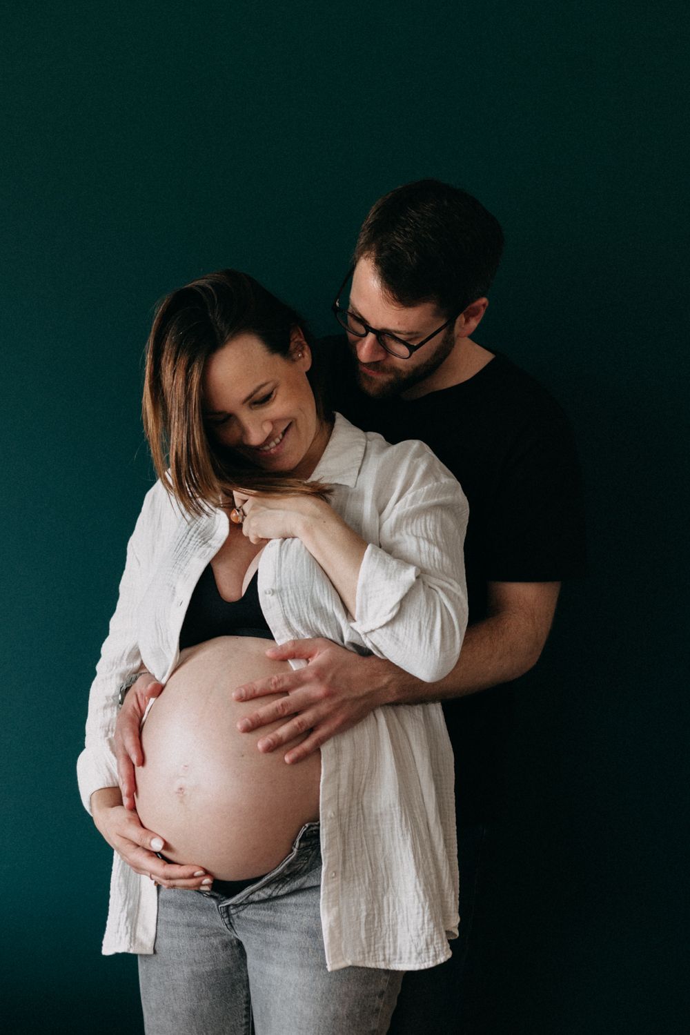 An intimate maternity portrait shows a tender moment between a couple against a dark green wall.