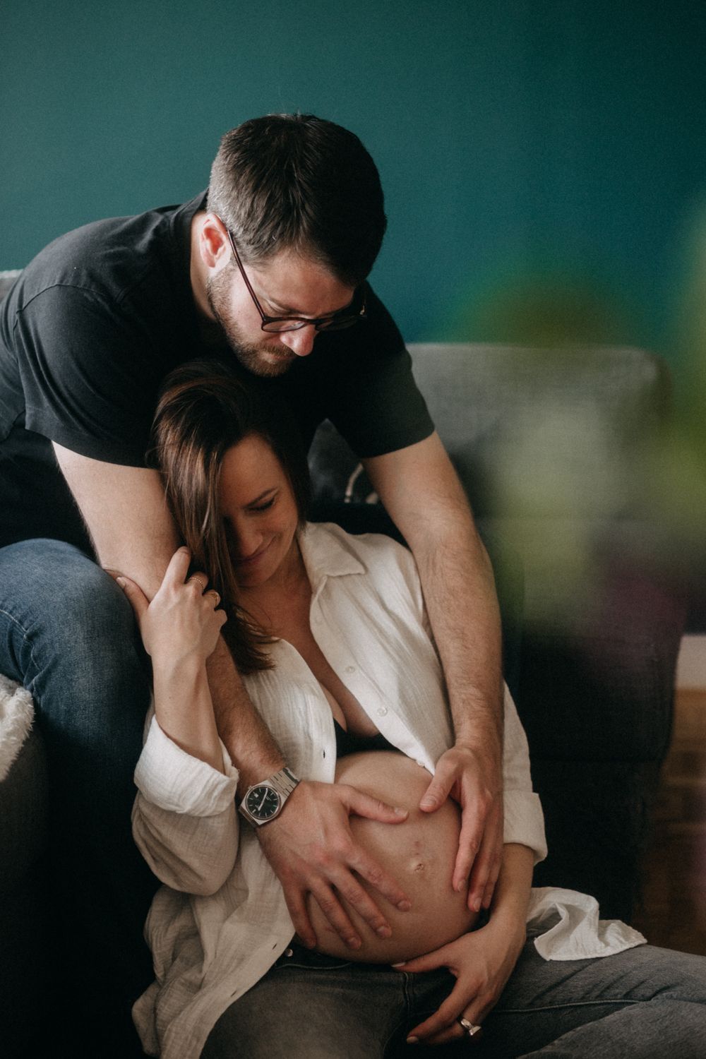 An expecting couple shares an intimate moment as they tenderly embrace and cradle a pregnant belly against a teal background.