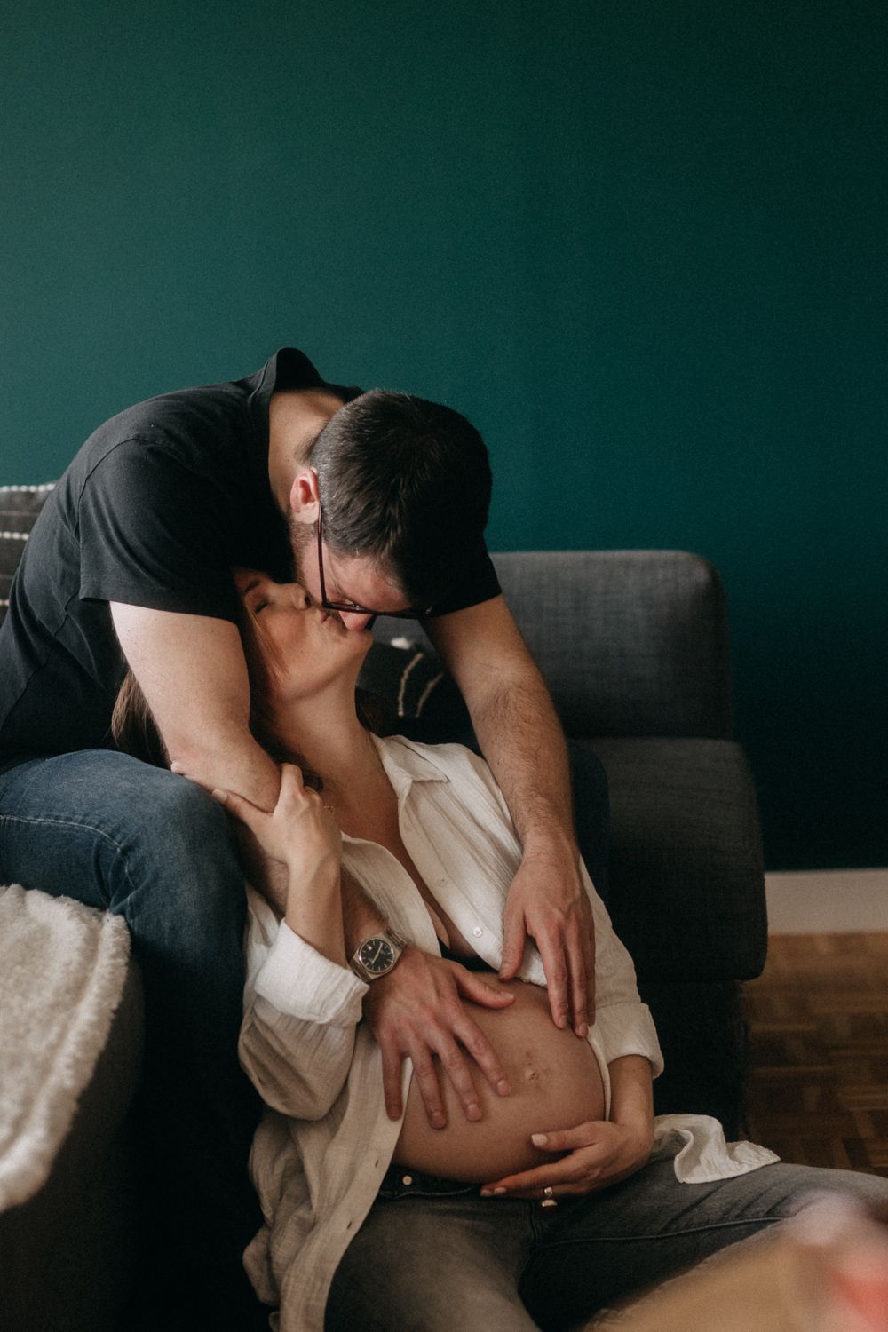 Intimate maternity photoshoot showing tender moment with couple on gray couch against dark teal wall.