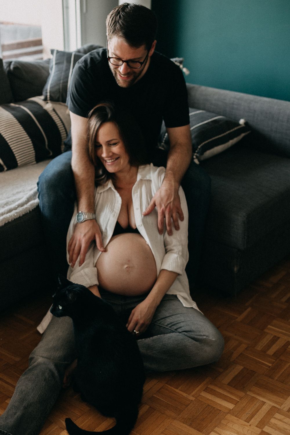 Intimate maternity photo showing expecting couple sharing tender moment on dark couch against teal wall.