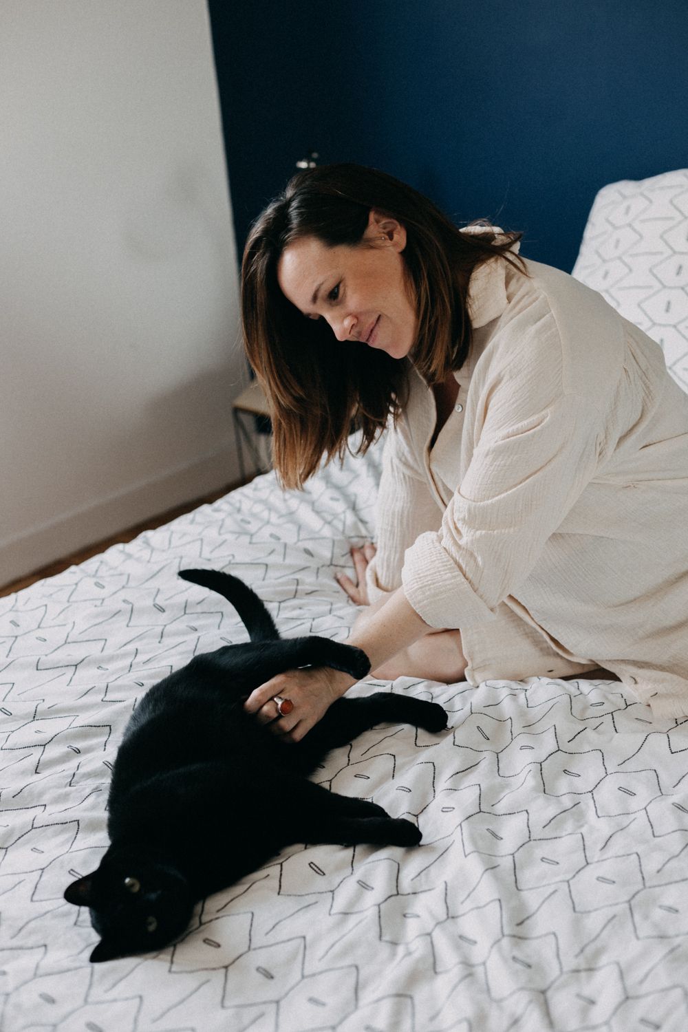 Woman in white robe sitting on patterned bedding while petting a black cat that is lying on the bed.