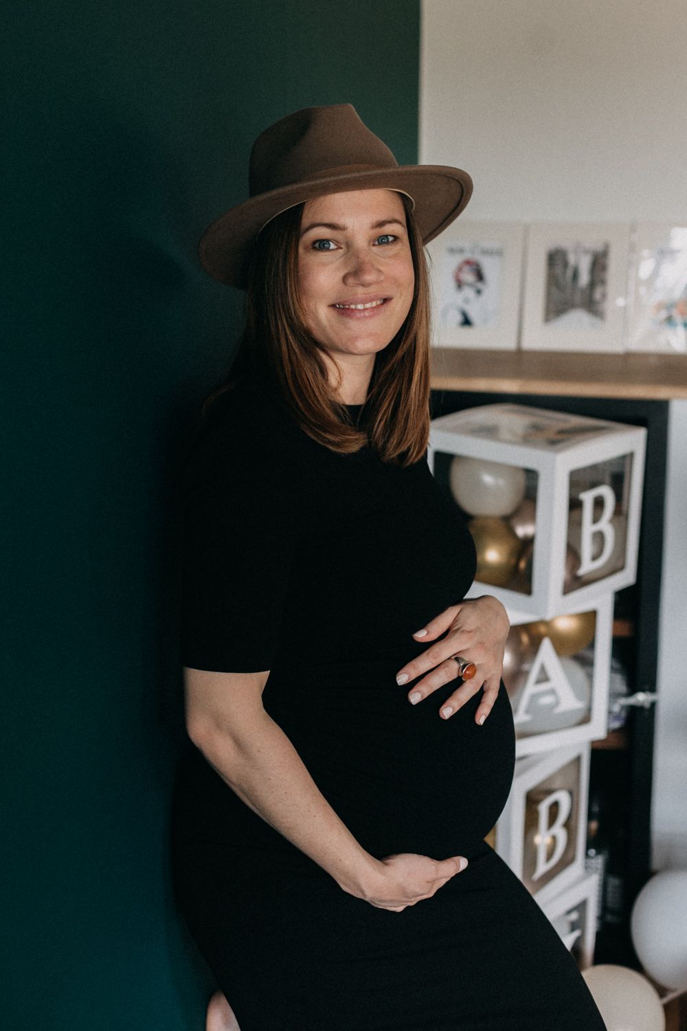 Expectant parent wearing brown hat and black dress poses against dark green wall with baby-themed decor in background.