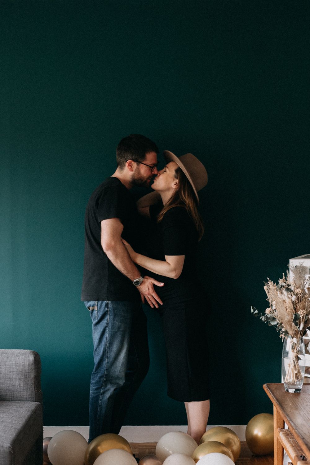 Couple in black outfits embrace against dark teal wall with gold and white balloons arranged along floor.