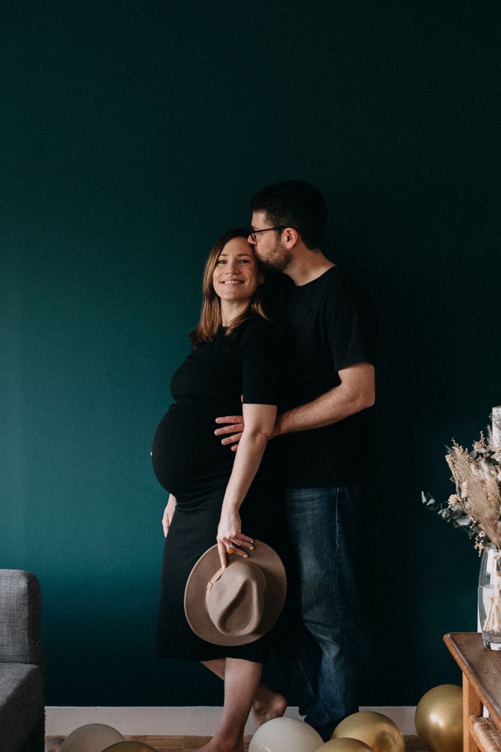 Couple shares intimate moment against dark teal wall with beige hat and balloons in maternity photoshoot.