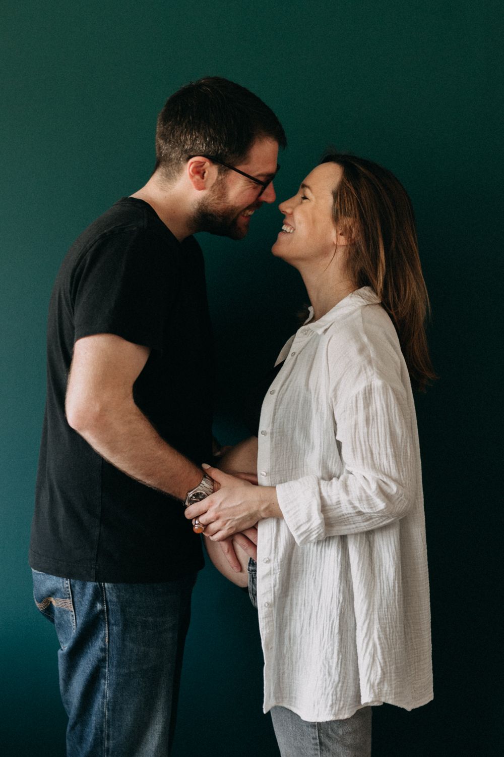 An intimate maternity portrait shows a couple touching foreheads against a dark green background.
